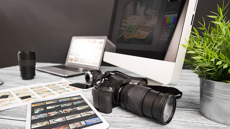 Máquina fotográfica, computador portátil, tablet com thumbnails, objetivas e provas de contacto sobre uma mesa de trabalho